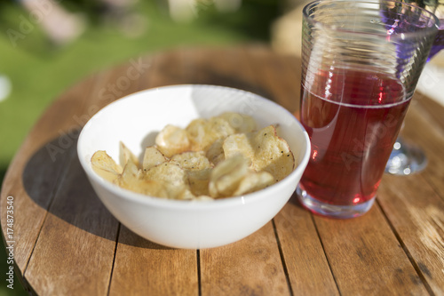 Single white bowl of crisps and a glass of juice outside on a wooden table in summer