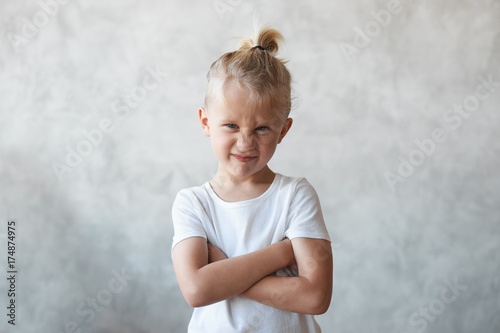 Tableau sur toile Angry stubborn Caucasian male kid with hair knot crossing arms on his chest and grimacing, expression unwillingness and protest towards doing something he doesn't want