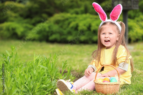 Cute little girl with basket of colorful eggs sitting on green grass in park. Easter hunt concept