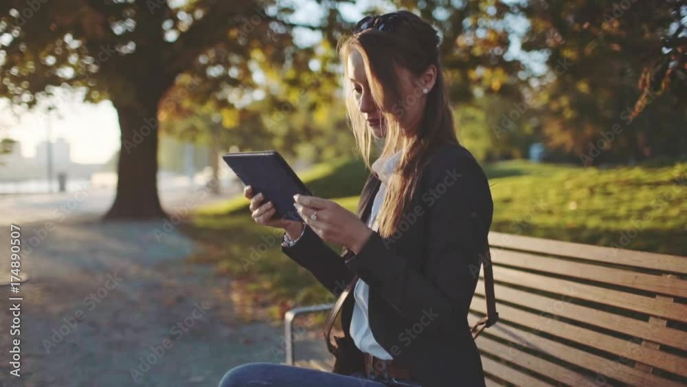 Young Businesswoman Using Digital Tablet, Sitting on the Bench in the Park in the Sunny Morning City. SLOW MOTION. STEADICAM Stabilized Shot. Business Woman Relaxing Outdoors. Lens Flare. Young Businesswoman Using Digital Tablet, Sitting on the Bench in the Park in the Sunny Morning City. SLOW MOTION. STEADICAM Stabilized Shot. Business Woman Relaxing Outdoors. Lens Flare.