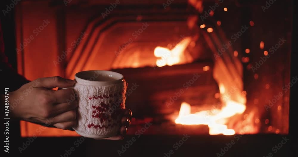 Woman Hands Holding Coffee or Tea Cup Against Cozy Fireplace in Winter. 4K SLOW MOTION 120 fps. Girl is having hot drink by the fire in the living room. Festive Winter Season Background. Woman Hands Holding Coffee or Tea Cup Against Cozy Fireplace in Winter. 4K SLOW MOTION 120 fps. Girl is having hot drink by the fire in the living room. Festive Winter Season Background.