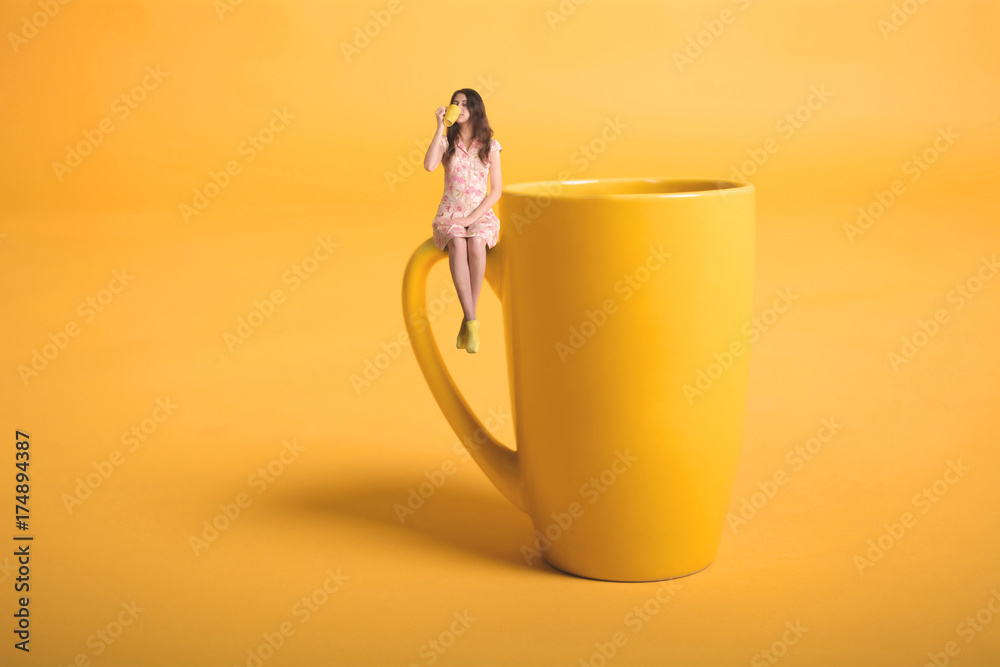 Creative surrealism design with miniature people. Girl with a cup. The girl in a pink dress sits on the mug. Yellow cup of tea or coffee. Young woman drinking tea. Mug on yellow background - obrazy, fototapety, plakaty 