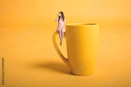 Creative surrealism design with miniature people. Girl with a cup. The girl in a pink dress sits on the mug. Yellow cup of tea or coffee. Young woman drinking tea. Mug on yellow background photo