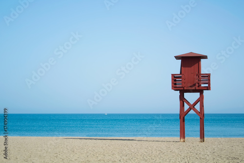Red wooden lifeguard tower on the abandoned beach at Benalmadena, Malaga province, Spain. Beautiful view of the sea and sandy beach on sunny summer day with copy space.