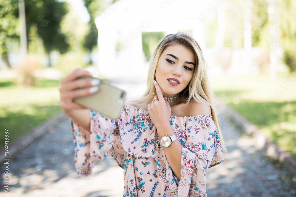 Young beautiful blonde girl relaxing in park, taking a selfie by mobile phone.