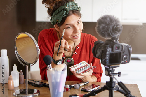 Portrait of happy young female beauty blogger using big palette and brush, applying eyeshadow while recording make up video tutorial, giving tips on how to accentuate color and shape of eyes photo