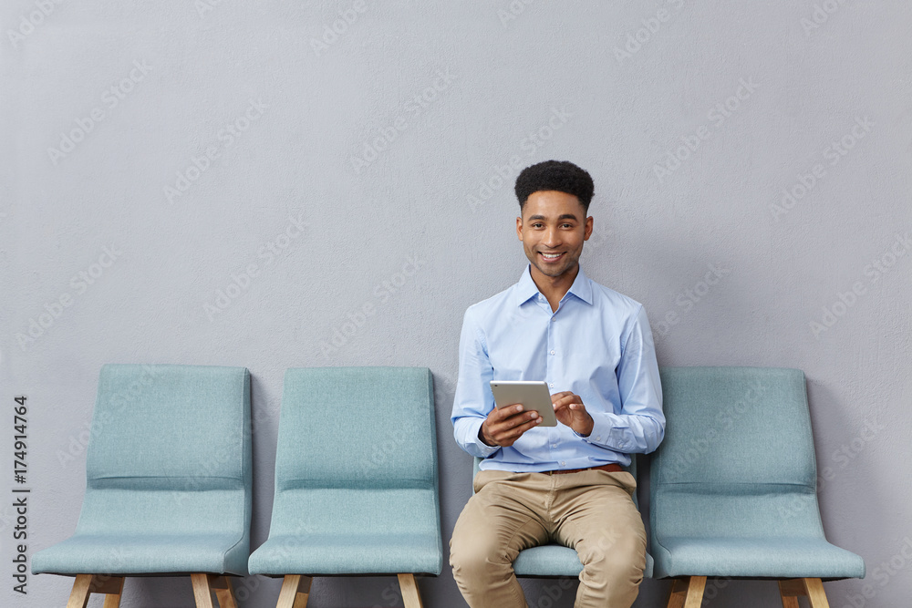 Horizontal portrait of mixed race male student sits at chair near classroom, being glad to pass exam successfully, holds tablet in hands, plays games online as waits for groupmates. Education concept