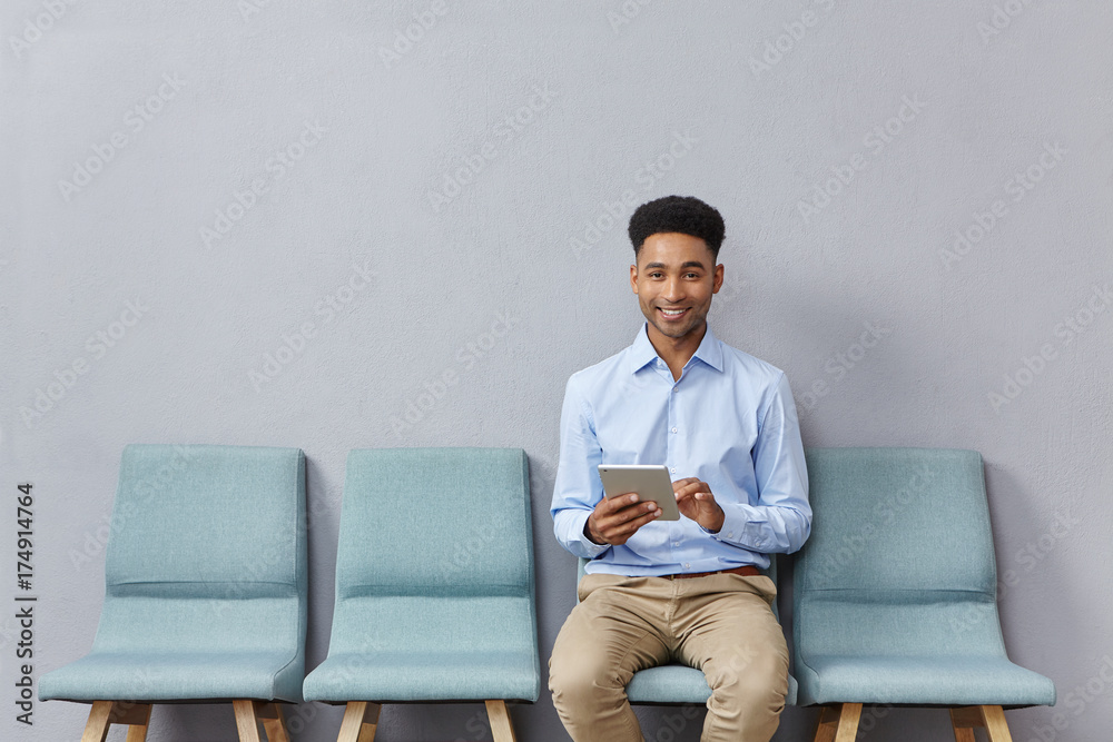 Horizontal portrait of mixed race male student sits at chair near classroom, being glad to pass exam successfully, holds tablet in hands, plays games online as waits for groupmates. Education concept Horizontal portrait of mixed race male student sits at chair near classroom, being glad to pass exam successfully, holds tablet in hands, plays games online as waits for groupmates. Education concept