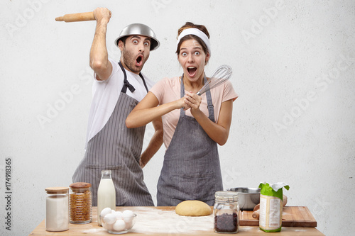 Picture of shocked professional restaurant workers have rivalry or competiton with each other, hold kitchen supplies, fight in funny manner, being surprised or scared to see chef, isolated over white