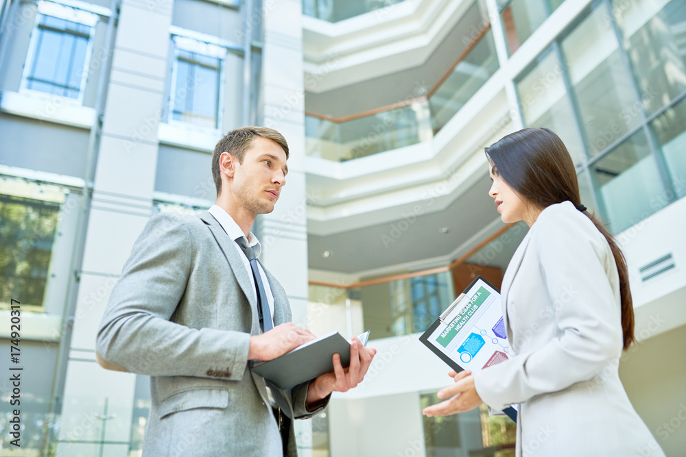 Low angle view of confident young manager presenting her point of view concerning start-up project while having informal meeting with colleague, interior of modern office lobby on background Low angle view of confident young manager presenting her point of view concerning start-up project while having informal meeting with colleague, interior of modern office lobby on background
