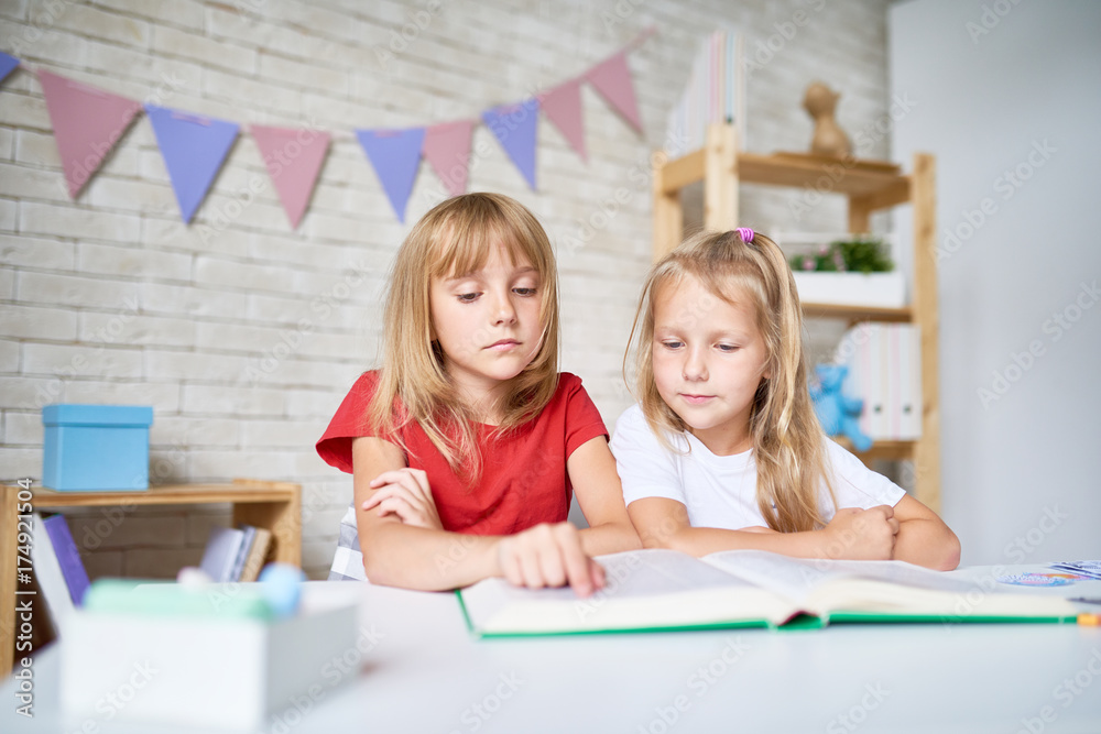 Portrait of two blonde girls reading big book together sitting at table in class of elementary school Portrait of two blonde girls reading big book together sitting at table in class of elementary school