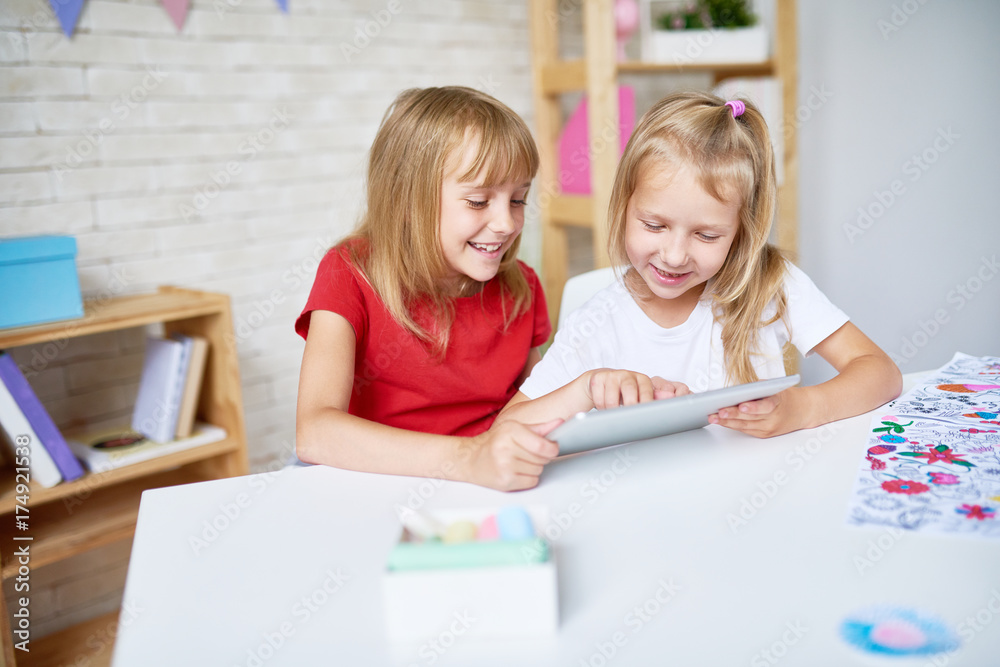 Portrait of two happy little sisters using digital tablet together and smiling sitting at desk in playroom Portrait of two happy little sisters using digital tablet together and smiling sitting at desk in playroom