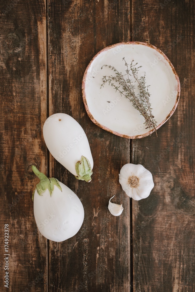 Close up top view of still life with white eggplants with garlic, dried herbs and ceramic handmade plate on rustic dark brown wooden table background. Vertical shot. Close up top view of still life with white eggplants with garlic, dried herbs and ceramic handmade plate on rustic dark brown wooden table background. Vertical shot.