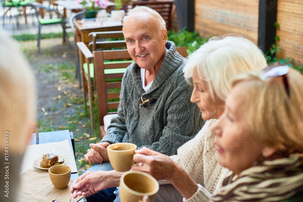 Portrait of handsome senior man with deep blue eyes listening to his friend with interest while having good time together at outdoor cafe, pretty women sitting next to him and enjoying fragrant coffee Portrait of handsome senior man with deep blue eyes listening to his friend with interest while having good time together at outdoor cafe, pretty women sitting next to him and enjoying fragrant coffee