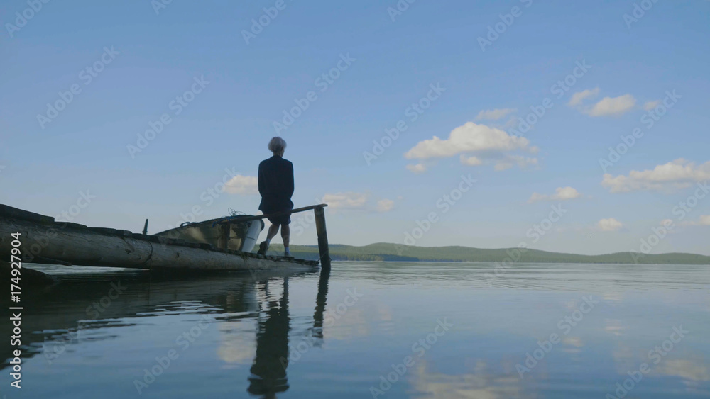 Naklejka premium Side view of a girl sitting on wooden pier on river bank. Side view of dreaming girl sitting on wooden pier on river bank. Beautiful woman with blond hair