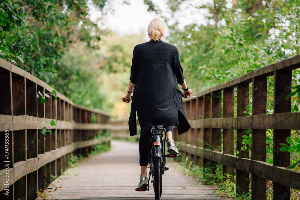 Young stylish blonde woman in black cardigan riding on bicycle on wooden road in park. Handsome girl having a ride. Back view
