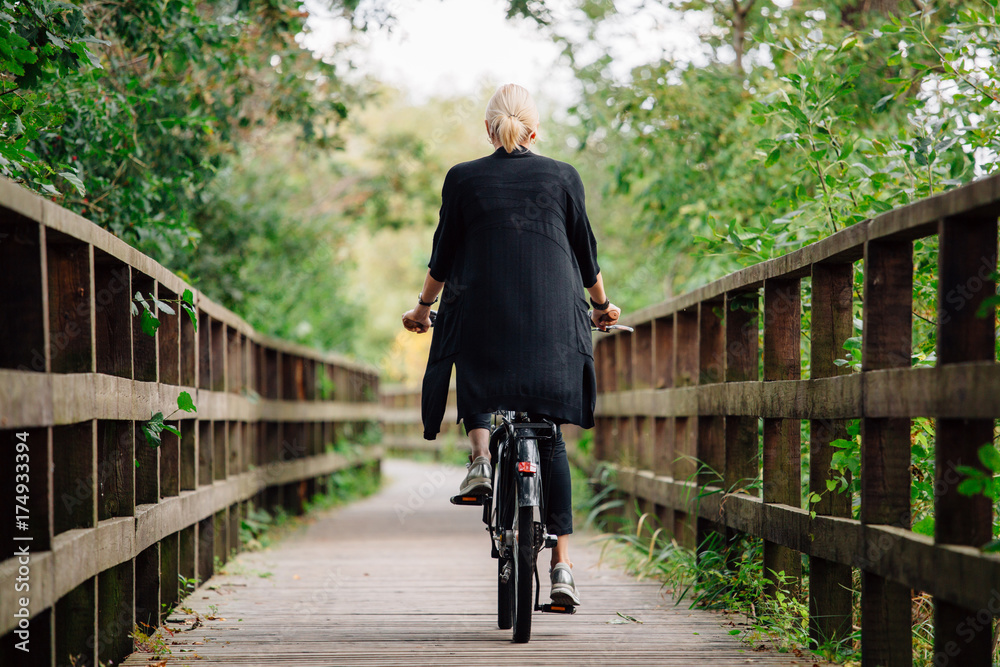 Young stylish blonde woman in black cardigan riding on bicycle on wooden road in park. Handsome girl having a ride. Back view