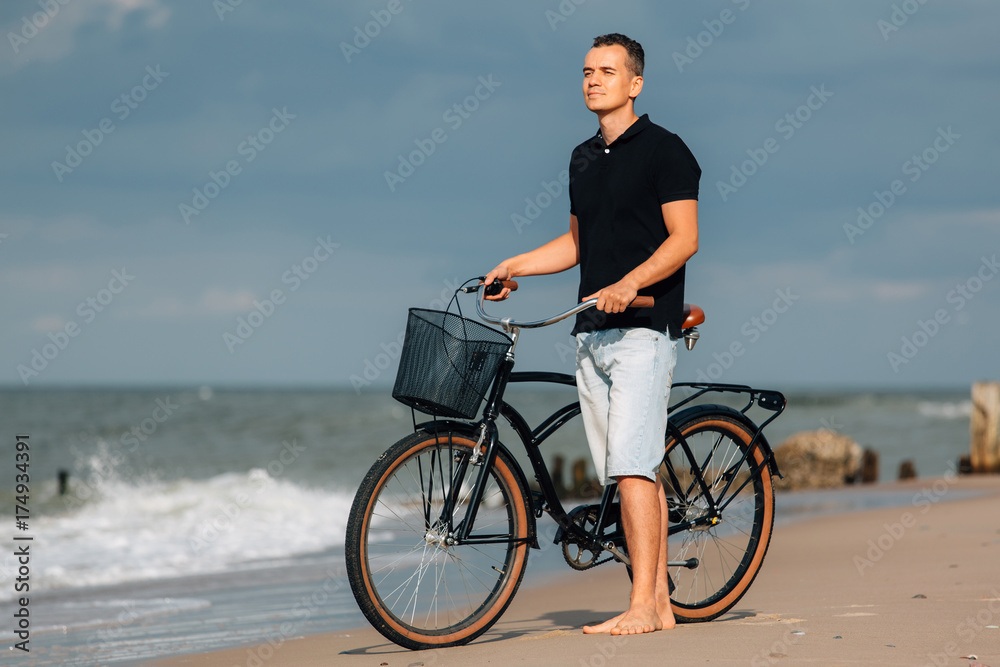 Pretty young man in casual walking on beach with retro bicycle. Handsome guy resting near sea