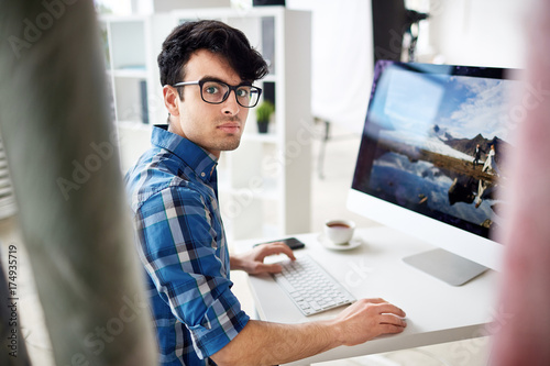 Serious young designer or retoucher sitting by workplace in front of computer and doing his work