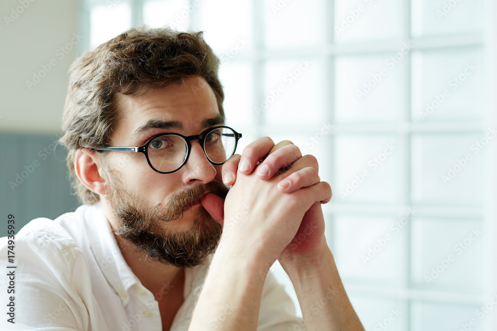 Young man in eyeglasses looking at camera while keeping his hands by mouth Young man in eyeglasses looking at camera while keeping his hands by mouth