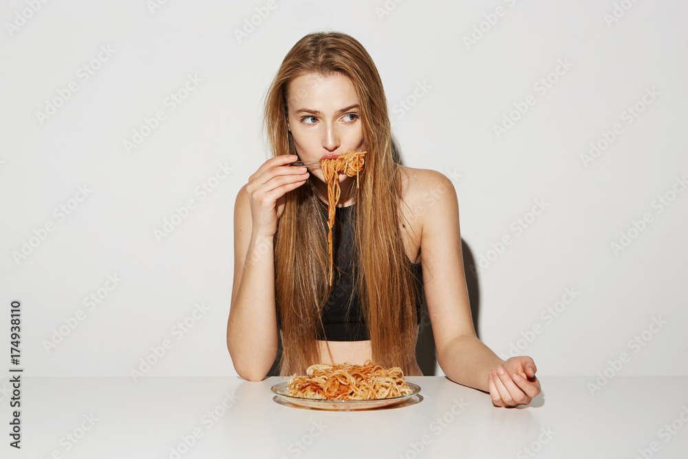 Close up portrait of sexy blonde girl with long hair sitting at table, eating spaghetti, looking aside with relaxed and flirty expression. Close up portrait of sexy blonde girl with long hair sitting at table, eating spaghetti, looking aside with relaxed and flirty expression.