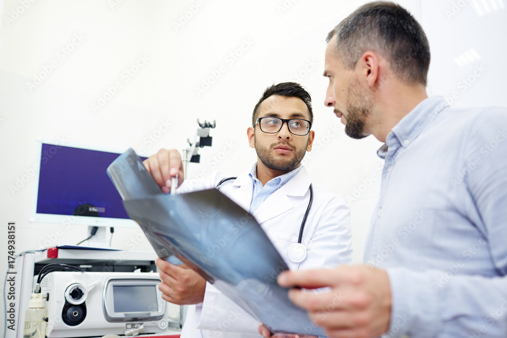 Obraz premium Highly professional radiologist analyzing X-ray images of middle-aged patient and giving necessary recommendations, interior of modern office on background