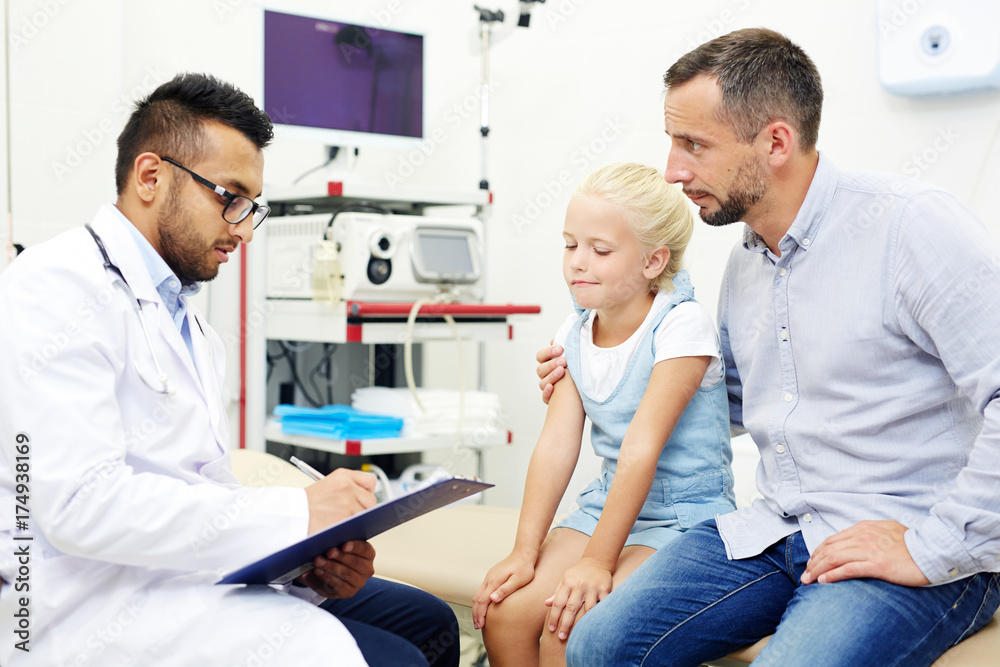 Pretty little girl and her handsome middle-aged father at pediatrician office: they looking at confident doctor with concentration while he filling in medical record