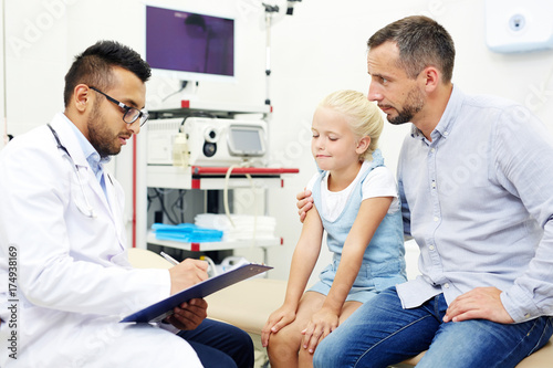 Pretty little girl and her handsome middle-aged father at pediatrician office: they looking at confident doctor with concentration while he filling in medical record