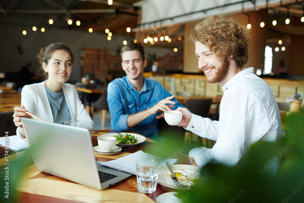 Young man with cup of tea looking at data on laptop display being pointed at by one of colleagues at meeting Young man with cup of tea looking at data on laptop display being pointed at by one of colleagues at meeting