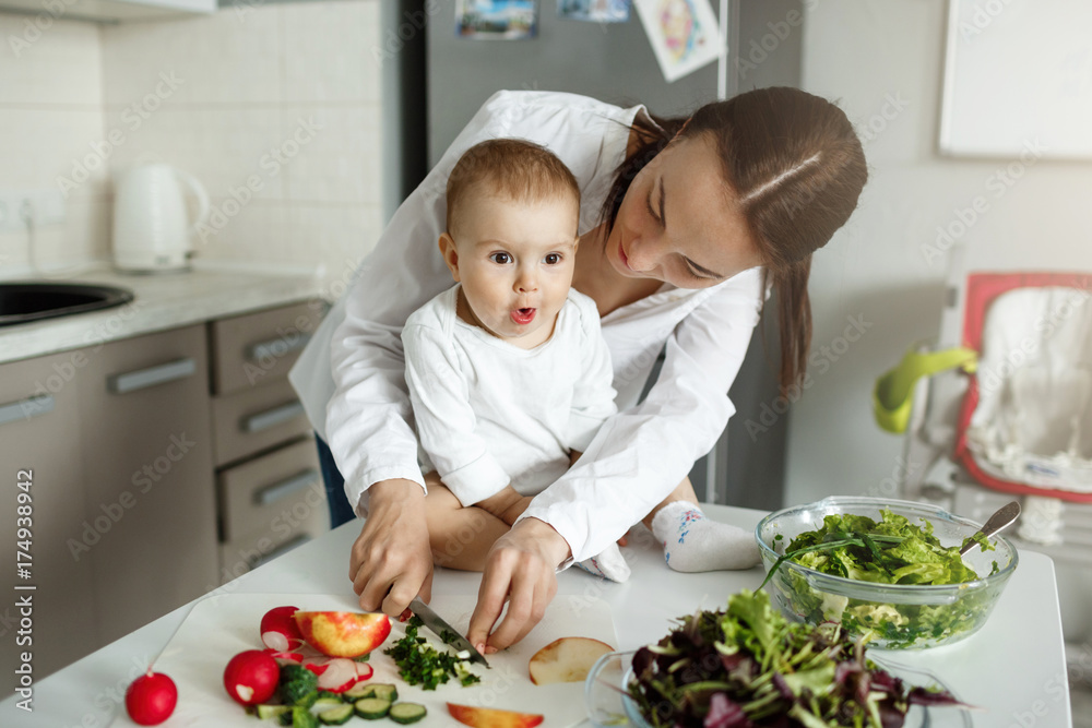 Beautiful dark haired mother cooking dinner in front of her adorable little son. Child looking aside with amezed expression. Beautiful dark haired mother cooking dinner in front of her adorable little son. Child looking aside with amezed expression.