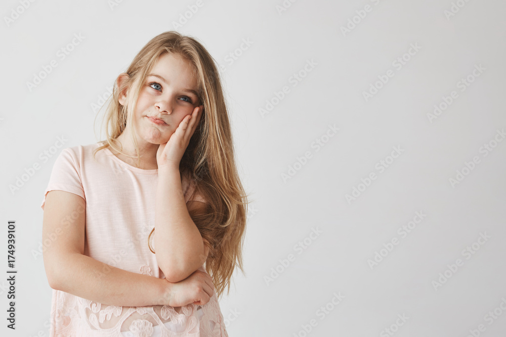 Little beautiful blonde girl with blue eyes squeezing face with hand, looking aside with frustrated expression after mom knows about bad marks at school. Little beautiful blonde girl with blue eyes squeezing face with hand, looking aside with frustrated expression after mom knows about bad marks at school.