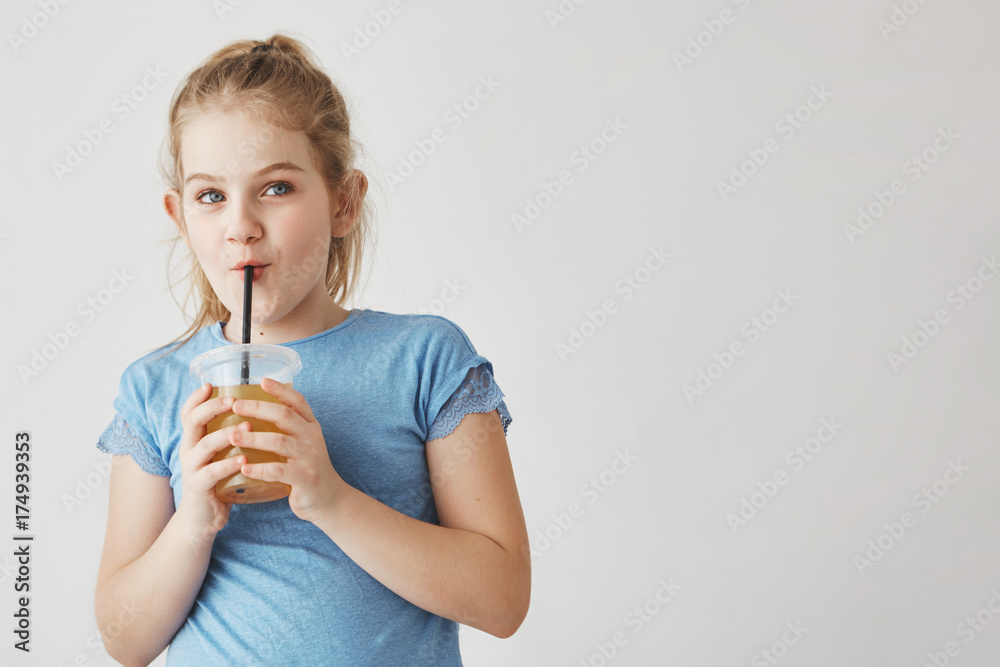 Close up of cute girl with light hair in tail hairstyle holding glass of milk in hands, drinking through straw and looking aside with funny face. Close up of cute girl with light hair in tail hairstyle holding glass of milk in hands, drinking through straw and looking aside with funny face.