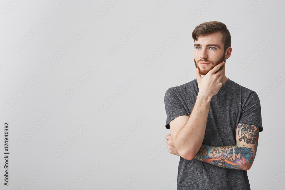 Close up of attractive young man with beard and tattoo on arm holding chin  with hand, looking aside with thoughtful expression. Stock Photo | Adobe  Stock