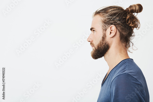 Close up of good-looking bearded hipster guy with hair in bun, in blue t-shirt standing in profile, looking aside, posing for photo.
