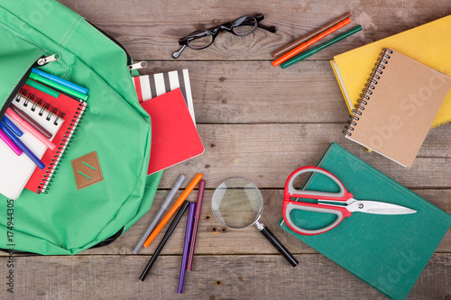 Backpack and school supplies: books, magnifying glass, notepad, felt-tip pens, eyeglasses, scissors on brown wooden table