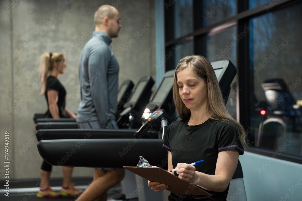 Woman writing on clipboard with people exercising on treadmill