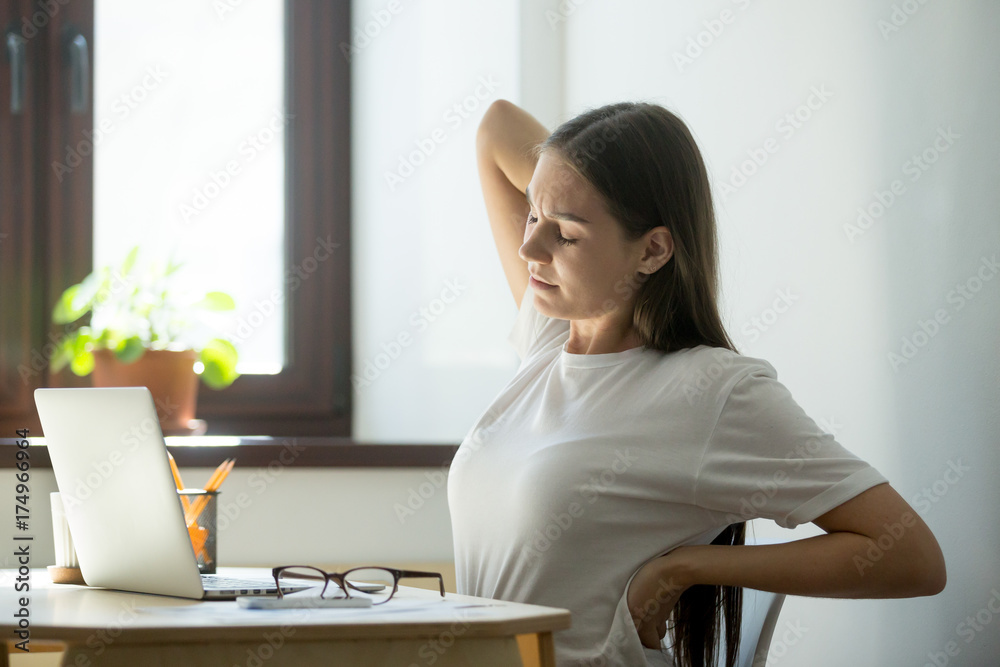 Female manager taking short break after long day of work on laptop computer. She took off her glasses, closed her eyes and stretching her aching back with a pained expression.