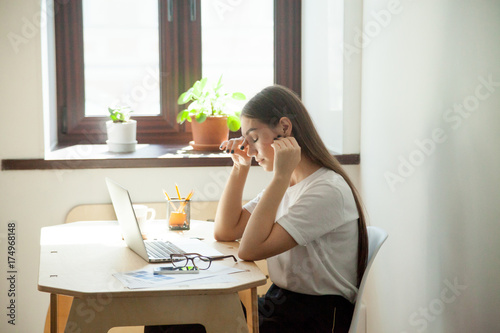 Female employee taking a minute break resting her aching eyes after computer work  having headache . Young tired woman sitting next to office window and pot plants feeling depressed