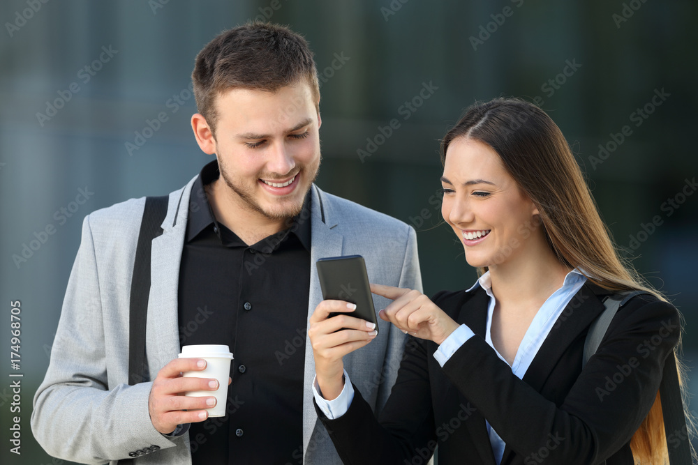 Front view of two happy executives talking about smart phone content standing outdoors on the street Front view of two happy executives talking about smart phone content standing outdoors on the street