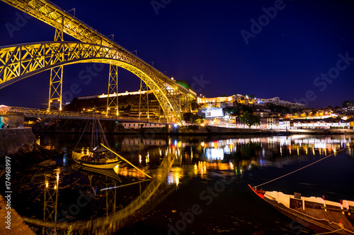 Iconic symbol of Oporto city. Scenic illuminated Dom Luis I bridge with boats reflecting on Douro River at night in Porto, Portugal's second largest city. Picturesque urban skyline. © bennymarty