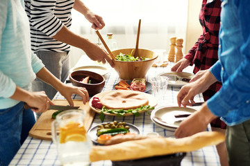 Mid-section group of young people preparing dinner for festive celebration standing at big table cutting vegetables for salad together