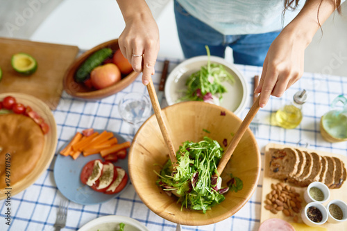 Closeup of female hands mixing green salad in wooden bowl at table with wholesome food while preparing family dinner