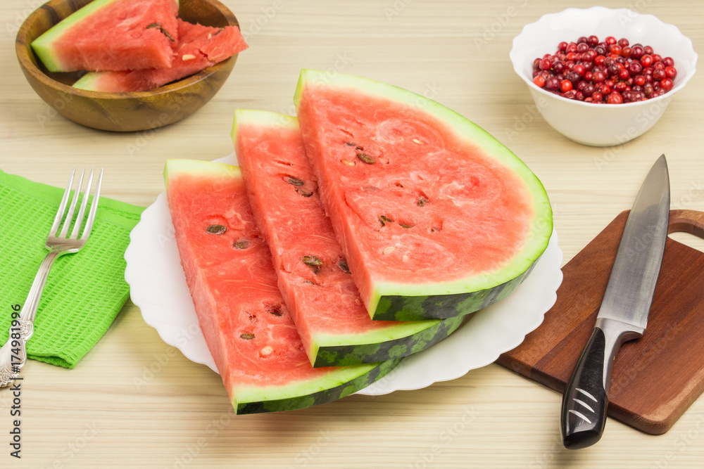 Delicious watermelon on a light wooden background. Three slices of berries ripe red watermelon on a white plate standing on the table with fork, knife. Green napkin, cranberries Delicious watermelon on a light wooden background. Three slices of berries ripe red watermelon on a white plate standing on the table with fork, knife. Green napkin, cranberries