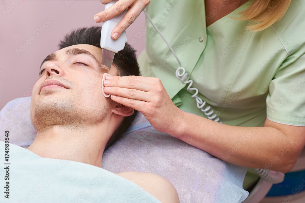 Hands using ultrasonic face scrubber. Cosmetologist working with young man. Hands using ultrasonic face scrubber. Cosmetologist working with young man.