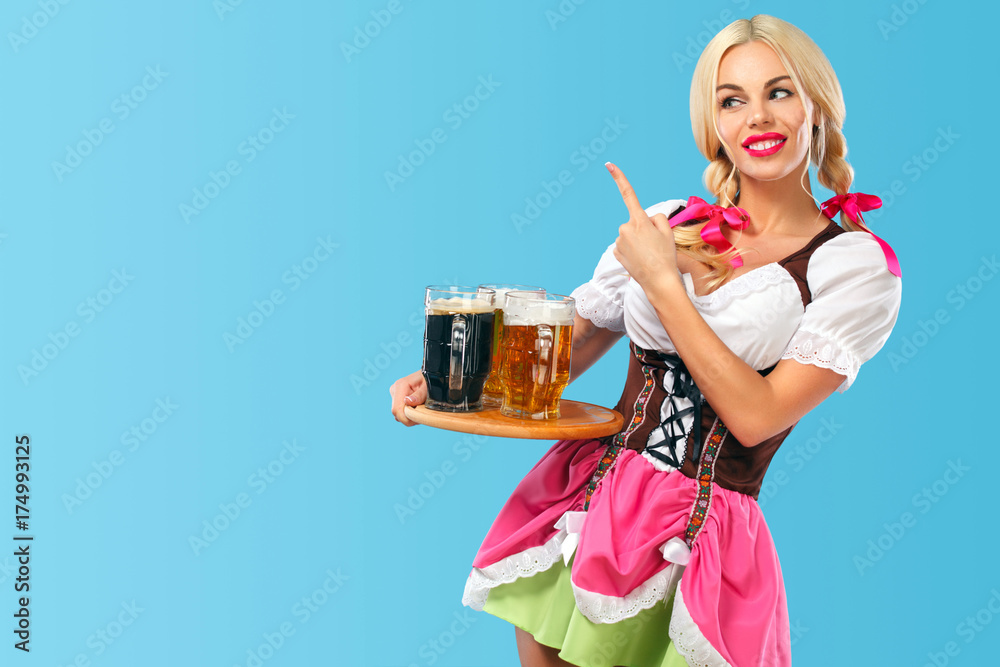 Young sexy Oktoberfest girl - waitress, wearing a traditional Bavarian dress, serving big beer mugs on blue background. Finger pointing on copy space Young sexy Oktoberfest girl - waitress, wearing a traditional Bavarian dress, serving big beer mugs on blue background. Finger pointing on copy space