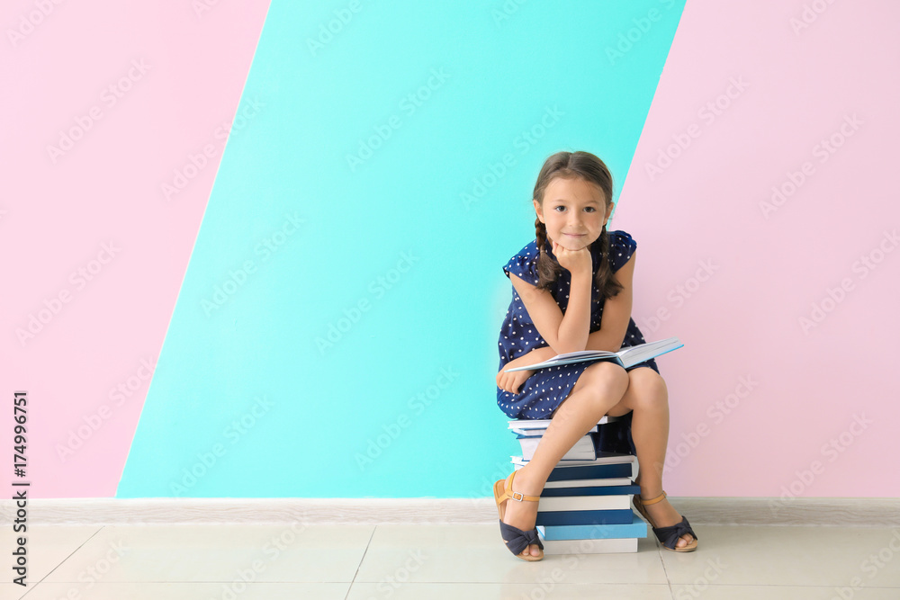Fototapeta premium Cute little girl sitting on pile of books near color wall