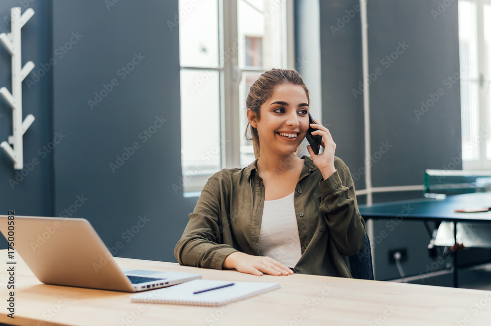 Portrait of a young woman taking a phone conversation sitting in a table at home office. Portrait of a young woman taking a phone conversation sitting in a table at home office.