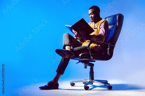 A black man is reading a book. A man is reading a book in a chair.