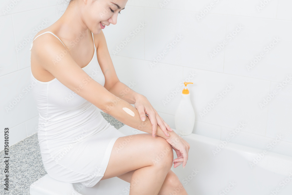 Young beautiful woman applying body lotion on her attractive legs in bathroom.