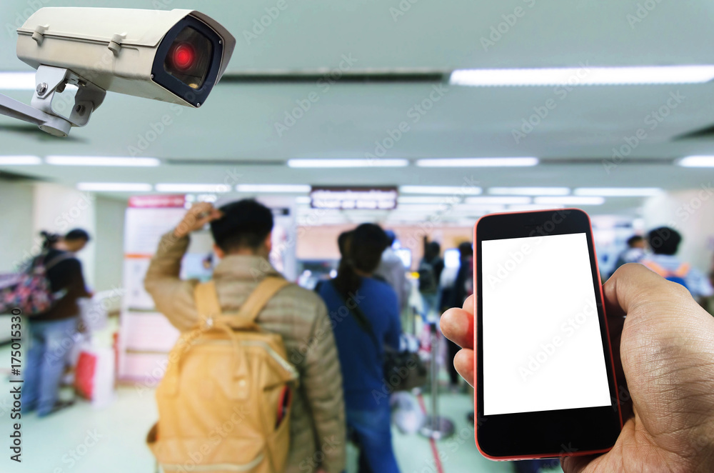 hand using smart phone monitoring and CCTV security indoor camera system operating in airport with people queue at immigration control background, surveillance security and safety technology concept
